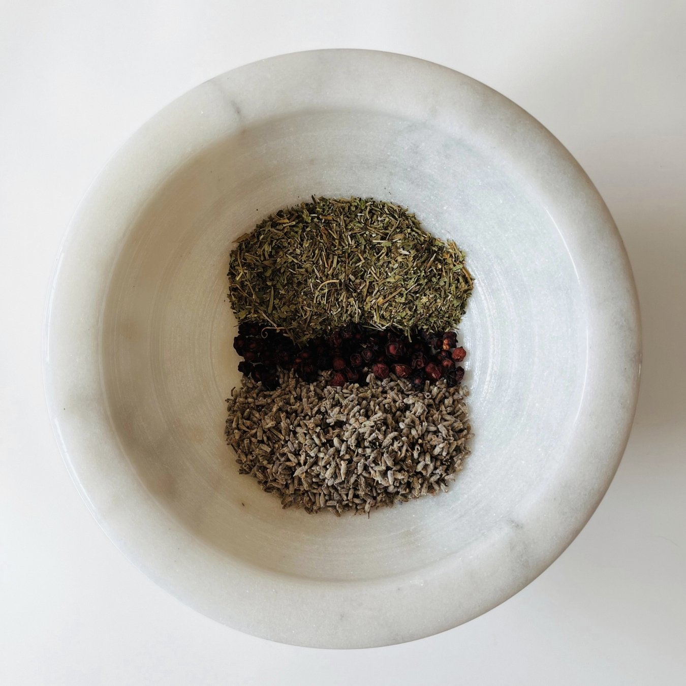 A variety of dried herbal tea leaves and ingredients, including gotu kola, berries, and lavender, arranged in a white marble bowl.